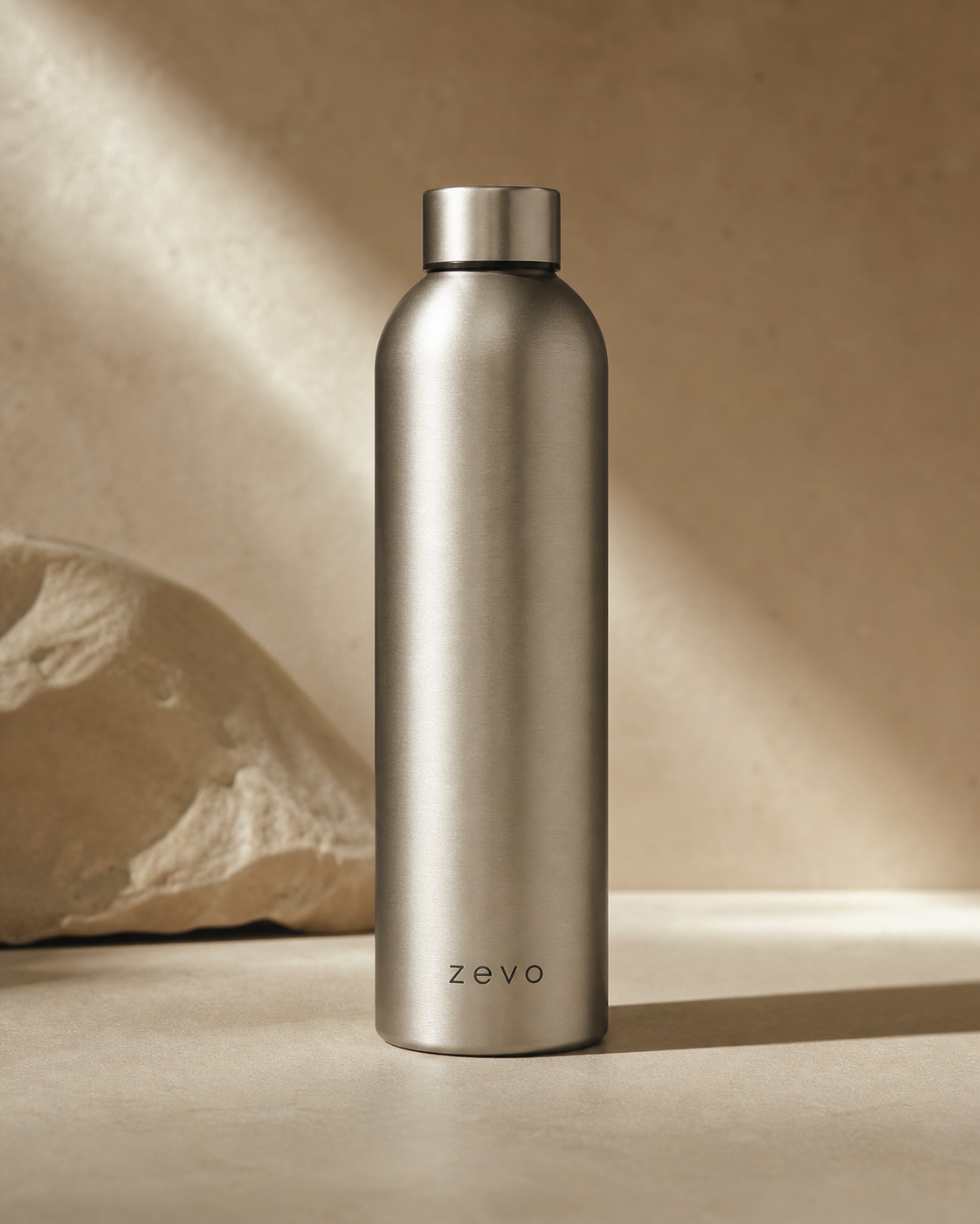 Zevo bottle in brushed stainless, photographed on warm sand backdrop with directional light